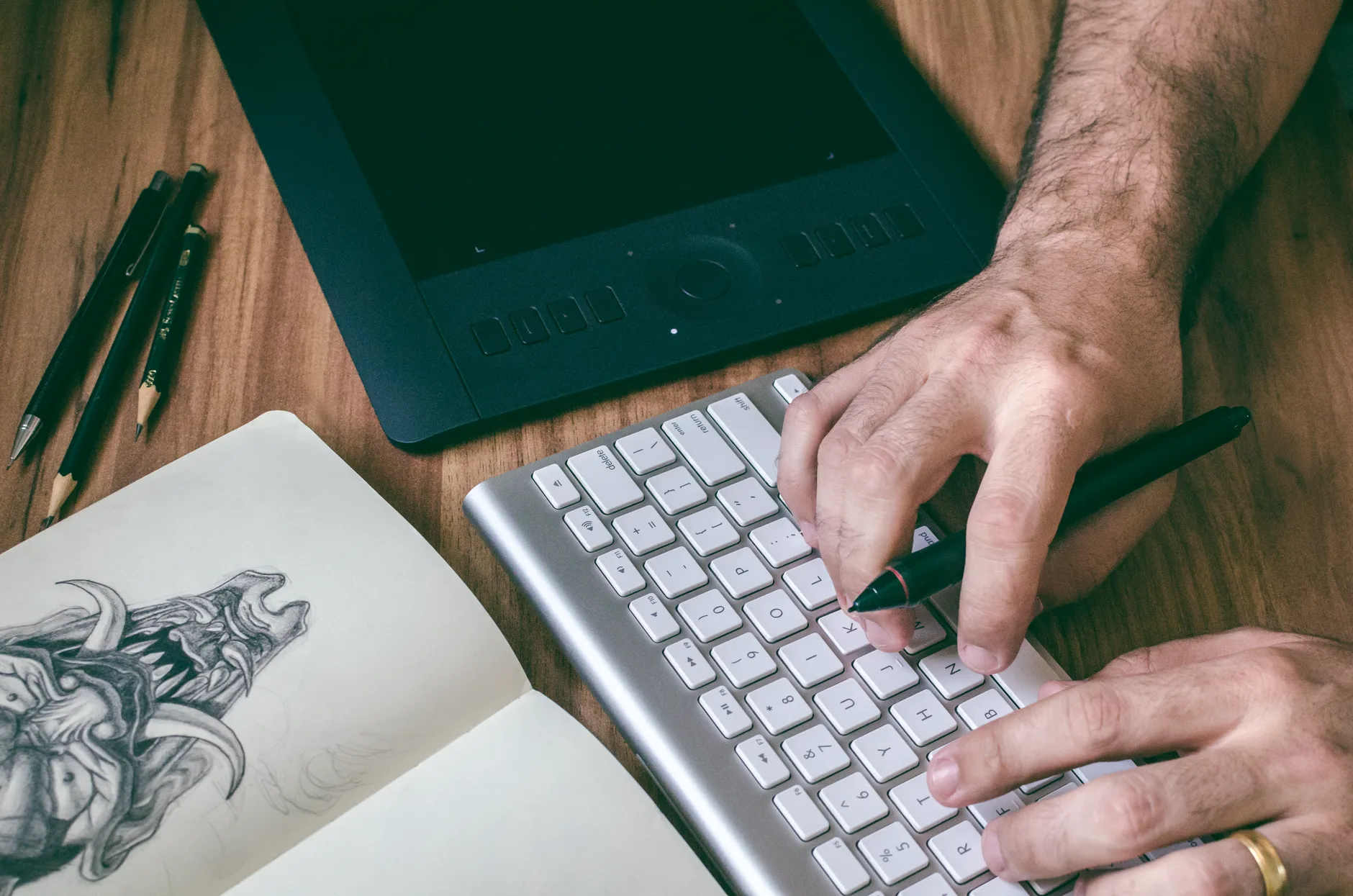 Hands working at a keyboard beside a pen tablet and a sketchbook with a drawing
