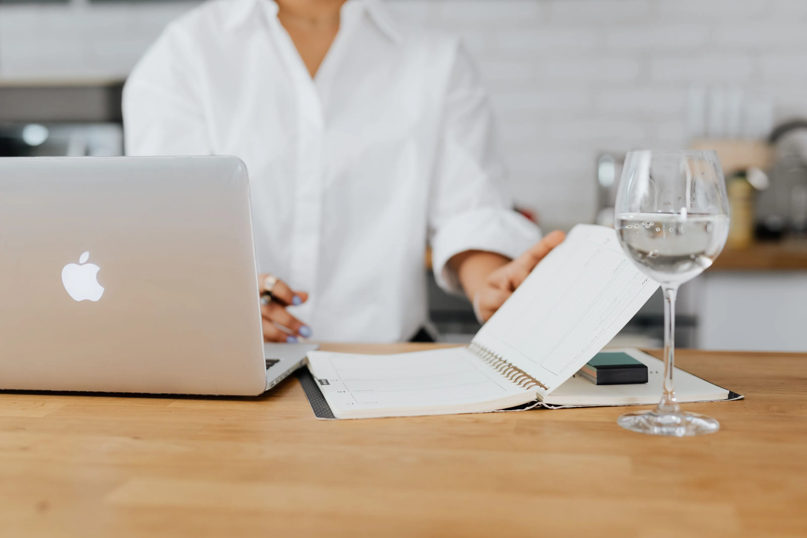 Woman in a white shirt working on a laptop with an open weekly planner and a glass of water on a wooden counter