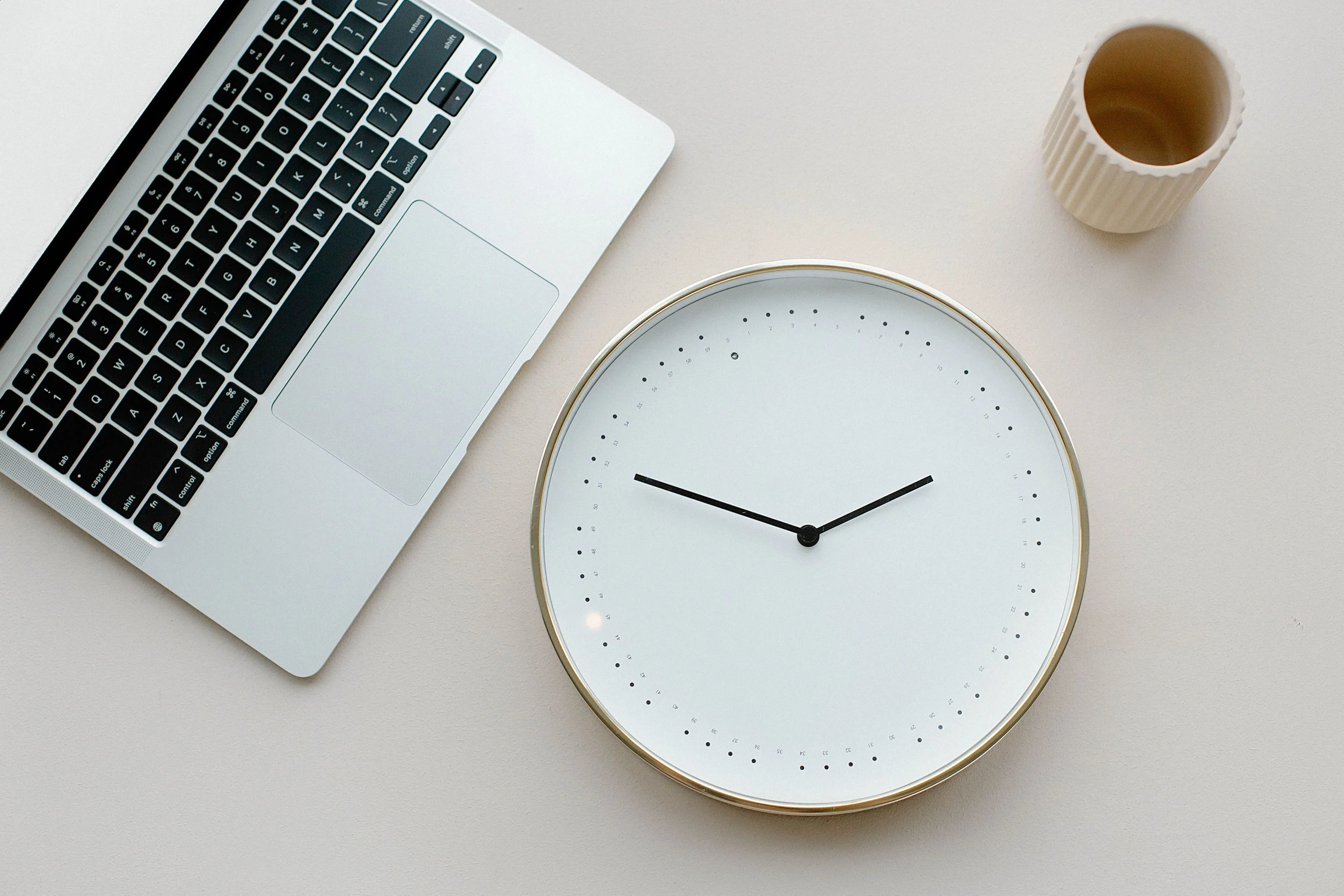 White wall clock beside an open laptop and a ceramic cup on a light desk, suggesting time blocking
