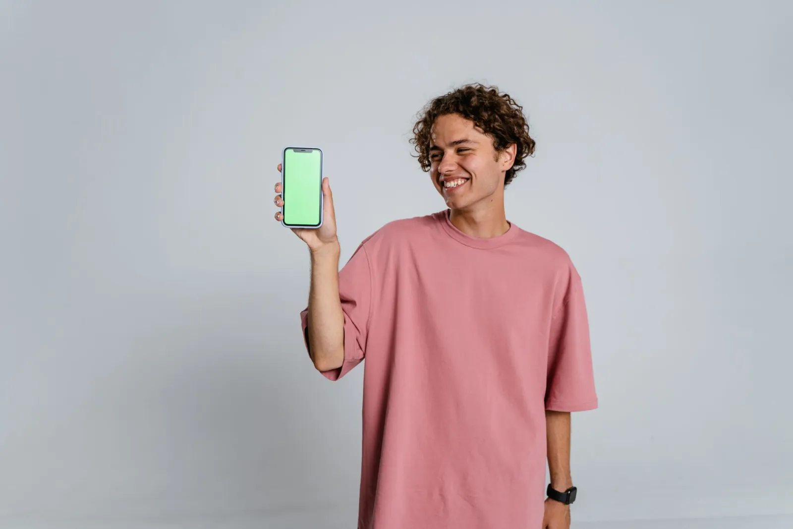 Smiling young man in a pink t-shirt holding a smartphone with a blank screen against a gray background.
