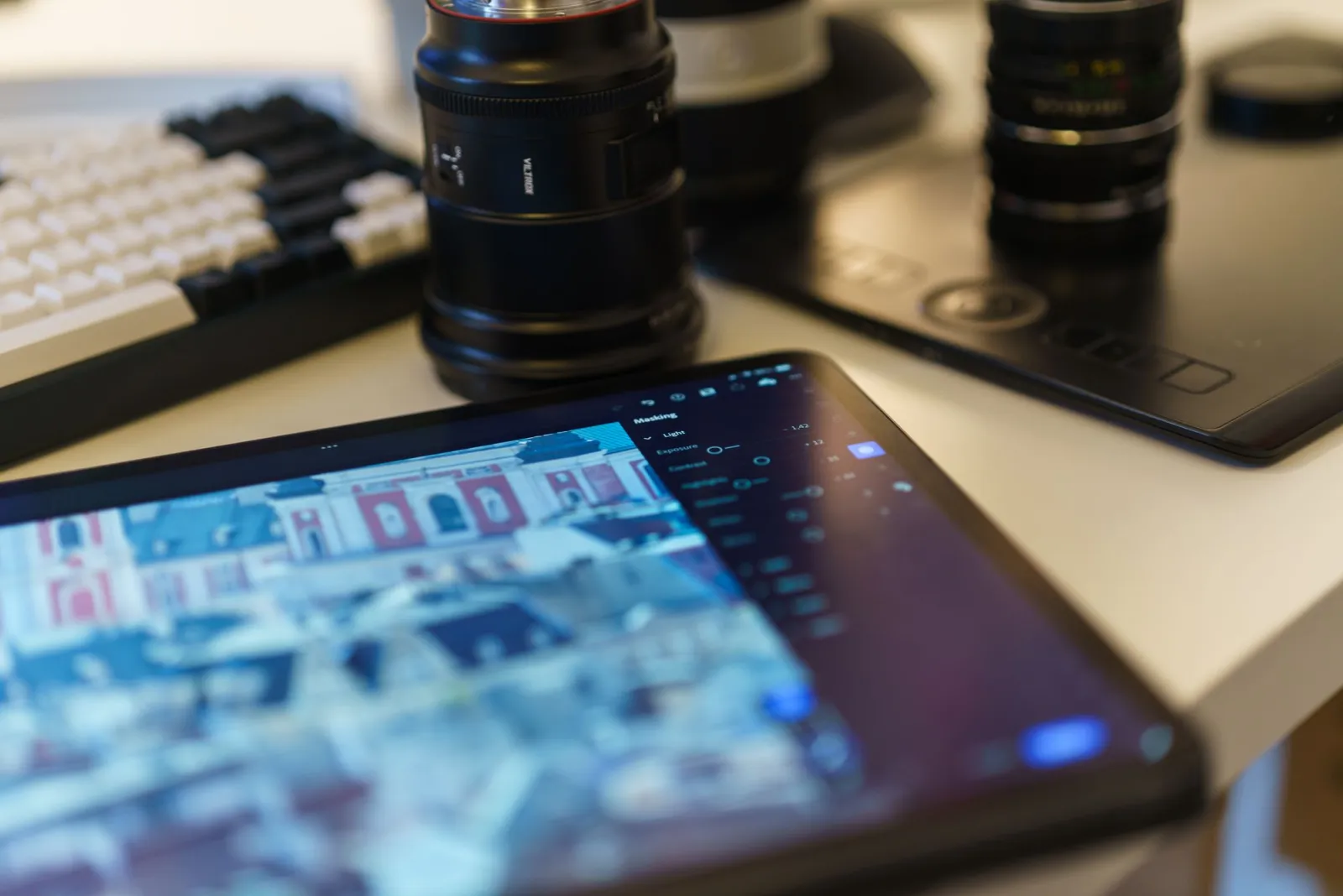 A modern desk workspace with a graphics tablet, camera lenses, and photo editing tools laid out for a post-processing session