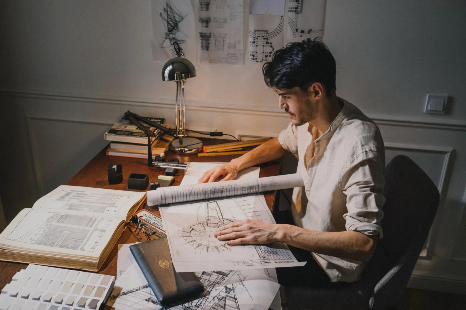 A designer working at a desk under warm lamp light surrounded by sketches and tools