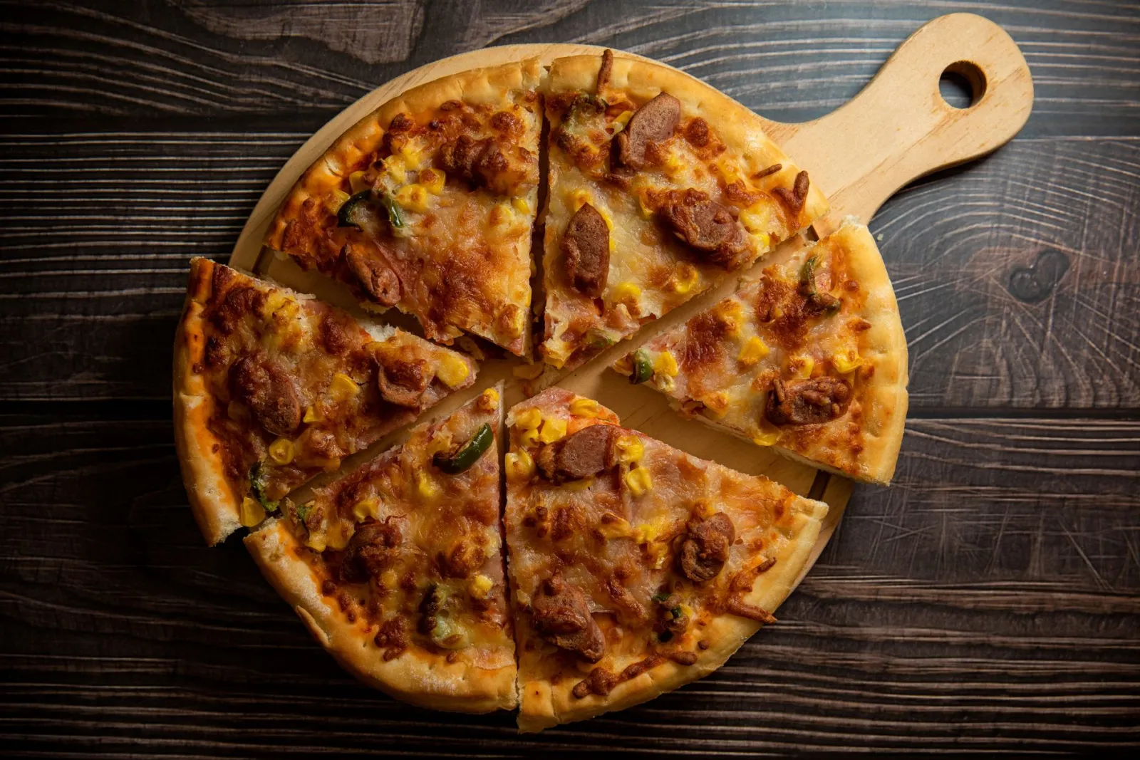 Appetizing sliced homemade pizza with sausage and corn on a rustic wooden board.