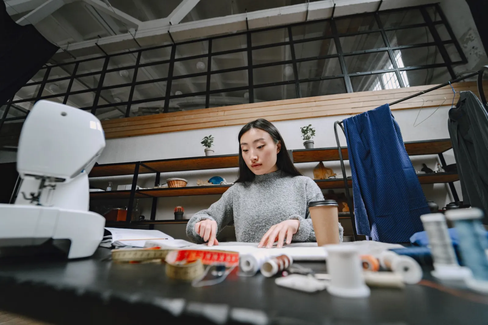A focused fashion designer works in a studio, surrounded by sewing tools, cloth, and a coffee cup